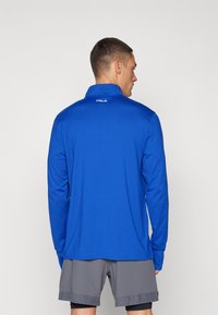 Langærmet blå atletisk skjorte i fugttransporterende stof med krave, prydet med et logo på ryggen, parret med grå shorts.