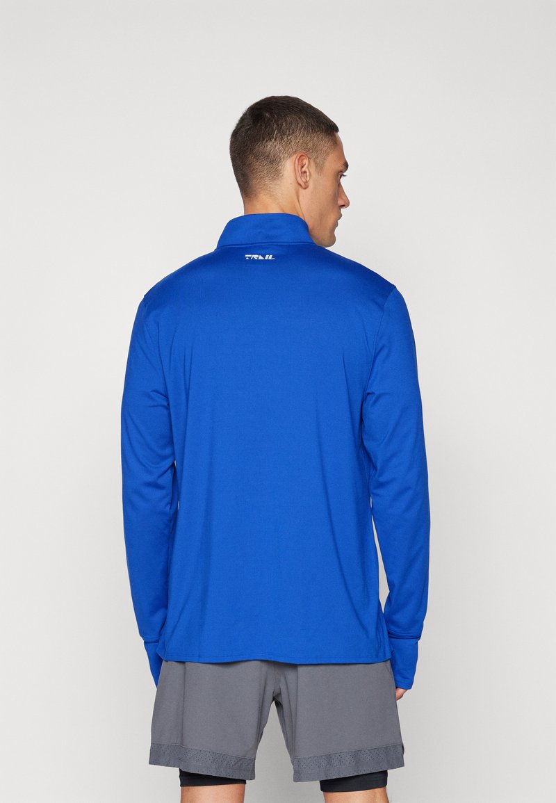 Camisa atlética de manga larga en tejido que absorbe la humedad, de color azul, con cuello, y con un logo en la parte posterior, combinada con pantalones cortos grises.