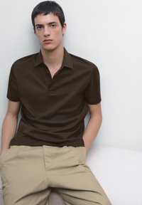 Mörkbrun polo-tröja med korta ärmar och en tre-knappars placket, i kombination med ljusbeiga byxor. Enkel, nedtonad design och passform.