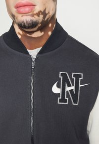 Mustat pommikaukalotakki, jossa on etuvetoketju, valkoinen 'N' -logo ja Nike-swoosh-logo, ribattu kaulus ja pehmeä kangasrakennus.