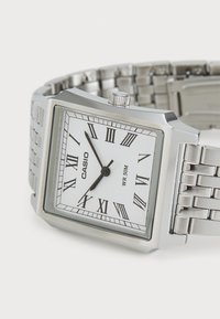 Montre Casio rectangulaire en argent avec un cadran blanc, des chiffres romains noirs et un bracelet en acier inoxydable. Résistante à l'eau, marquée WR-50M.
