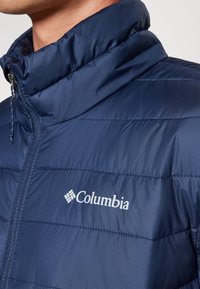 Tamsiai mėlyna izoliuota striukė su aukštu apykakle, siuvinėjimu ir priekiniais užtrauktukais. Ant krūtinės matomas baltas "Columbia" logo.
