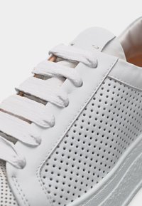 Λευκό δερμάτινο αθλητικό παπούτσι με χοντρά λευκά κορδόνια και διάτρητη πλαϊνή επιφάνεια, δείχνοντας κοντινή λεπτομέρεια της υφής και της ραφής.