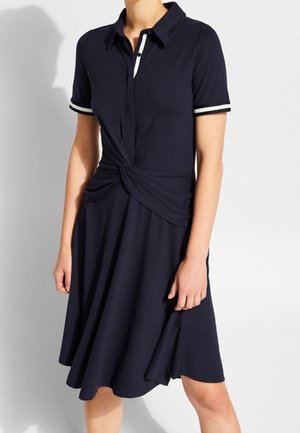 Femme portant une robe bleu marine à manches courtes avec un bord blanc au col et aux manches et un détail de nœud torsadé à la taille.