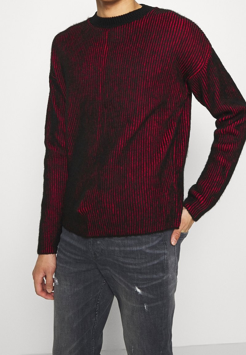 Pull rayé noir et rouge avec un col rond et des manches longues, caractérisé par une texture douce et un détail côtelé. Porté avec un jean gris.