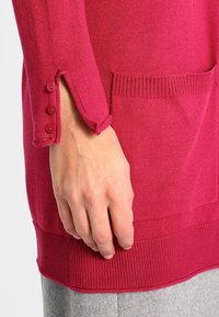 Suéter de punto en fucsia con dobladillo acanalado, que cuenta con un bolsillo y tres botones decorativos en el puño. Textura suave, diseño casual.