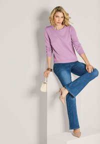 Frau in lila Pullover und blauen Jeans, die auf einem Bein balanciert und eine kleine beige Handtasche vor einem schlichten hellen Hintergrund hält.