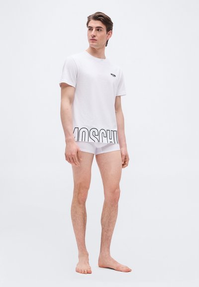 Νέος άνδρας όρθιος, ξυπόλητος, φοράει λευκό T-shirt Moschino και λευκά μπόξερ, μπροστά σε ένα απλό φωτεινό φόντο.