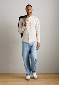 Man in een beige shirt en lichtblauwe jeans die een zwart jack over zijn schouder houdt, staande op een houten vloer tegen een effen muur.
