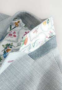 Grauer strukturierter Blazer mit einem floral gemusterten Satinfutter, ausgestattet mit einem grauen Wollkragenakzent. Klare Linien und maßgeschneiderte Gestaltung.