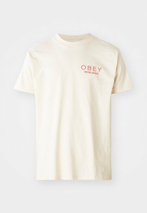 T-shirt en coton couleur crème avec des manches courtes, col rond et texte graphique rouge "OBEY MCMLXXXIX" imprimé sur le devant.