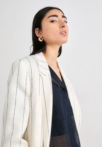 Blazer blanco con rayas negras, superpuesto a una blusa oscura y transparente. Pendientes de aro dorados añaden un acento metálico. Detalles de tela texturizada visibles.