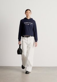 Námořnicky modrá krátká mikina s bílým logem, doplněná volnými bílými kalhotami a černými botami. Drží malou černou kabelku, na světlém pozadí.