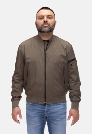 Uomo con capelli corti scuri e barba che indossa una giacca bomber marrone con zip e jeans blu, in piedi davanti a uno sfondo chiaro e semplice.
