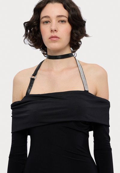 Robe noire sans épaule avec des manches longues et une silhouette cintrée. Présente des bretelles noires et argentées avec un accent de matériel décoratif.