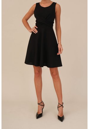 Femme portant une robe noire sans manches, arrivant au genou, et des chaussures à talons aiguilles noires avec des sangles à la cheville, se tenant avec une main sur la hanche.