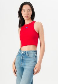 Rotes geripptes Crop-Top mit Rundhalsausschnitt und blauem Logo-Tag, kombiniert mit hochtaillierten hellblauen Jeans, vor einem schlichten weißen Hintergrund.
