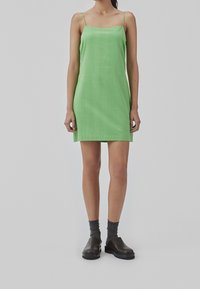 Robe nuisette en lin vert avec des bretelles fines, coupe droite, longueur genoux. Portée avec des sabots foncés et des chaussettes grises, se tenant devant un arrière-plan neutre.