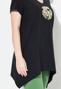 Zwarte tuniek met korte mouwen en een v-hals, voorzien van een abstract geborduurd ontwerp. De tuniek heeft een hi-lo zoom en wordt gecombineerd met groene leggings.