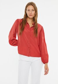 Camisa roja de manga larga con botones en la parte delantera, hombros con pliegues y un corte relajado. Hecha de una tela suave, combinada con pantalones blancos.