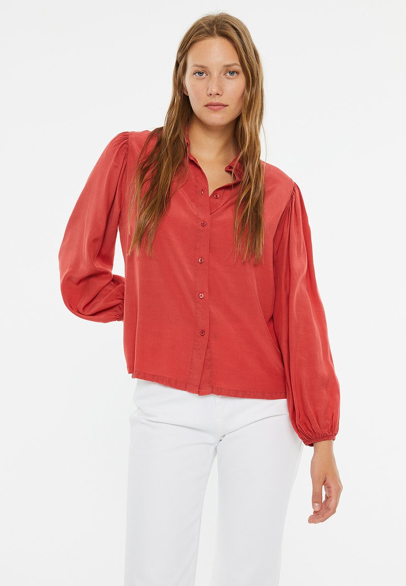 Camisa roja de manga larga con botones en la parte delantera, hombros con pliegues y un corte relajado. Hecha de una tela suave, combinada con pantalones blancos.