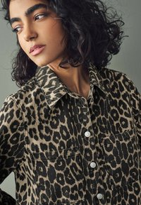 Cămașă din denim cu imprimeu de leopard, cu guler ascuțit, scheme de culori negru și bej, buzunare frontale și nasturi metalici. Material texturat.