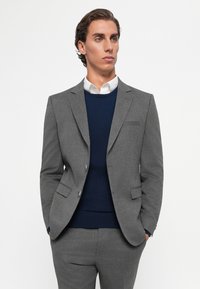 Mann in einem grauen Anzug mit einem marineblauen Pullover und weißem Hemd, steht mit den Händen in den Taschen und schaut etwas nach rechts.