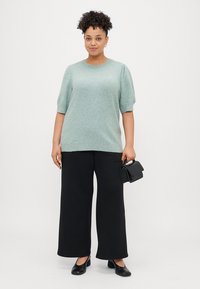 Ανοιχτό πράσινο πλεκτό πουλόβερ με κοντά φουσκωτά μανίκια, συνδυασμένο με φαρδιές μαύρες παντελόνες. Μια μικρή μαύρη τσάντα χεριού ολοκληρώνει την εμφάνιση.