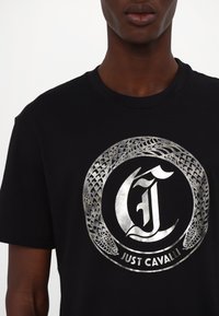 Juoda medvilninė marškinėliai su sidabro spalvos logotipo spausdinimu, kuriame pavaizduota įsivijusi gyvatė ir apskritimas, taip pat tekstas "JUST CAVALLI."