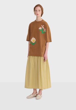 Frau trägt ein braunes, übergroßes T-Shirt mit pixeligen Blumendesigns, einen langen gelben Rock und beige Schlupfschuhe, steht vor weißem Hintergrund.