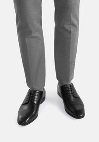 Melnas ādas formālās kurpes ar gludu apdari, apaļiem pirkstiem un savelkamu dizainu, apvienotas ar gaiši pelēkiem piegrieztiem biksēm.