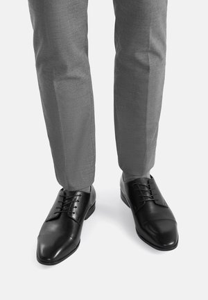 Juodos odinės batų su sklandžiu paviršiumi, apvaliomis pirštų dalimis ir raištelių dizainu, derinamos su šviesiai pilkomis pritaikytomis kelnėmis.