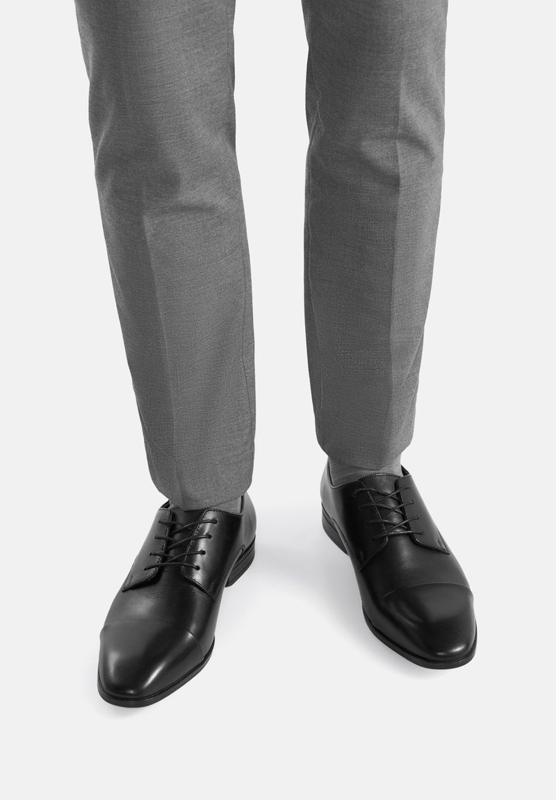 Melnas ādas formālās kurpes ar gludu apdari, apaļiem pirkstiem un savelkamu dizainu, apvienotas ar gaiši pelēkiem piegrieztiem biksēm.