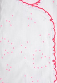Tessuto bianco con una texture a righe, caratterizzato da piccoli motivi a cuore rosa. Rifinito con volant rosa brillante e fissato da bottoni bianchi.