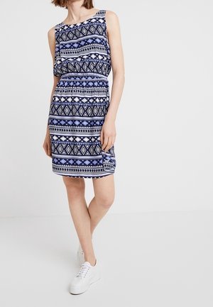 Robe sans manches marine avec un motif géométrique blanc et bleu clair, taille cintrée et longueur au genou, associée à des baskets blanches.