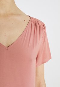 Różowa bluzka z dekoltem w serek i krótkimi rękawami, z marszczeniami na ramionach i gładką, miękką fakturą materiału.