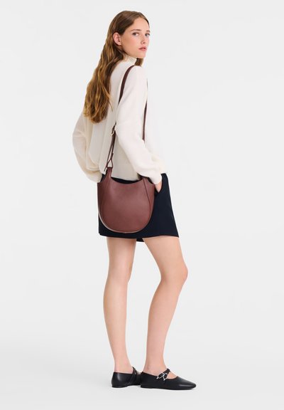 Brown leather handbag with a rounded shape, worn crossbody; paired with a light sweater and black skirt, featuring flat black shoes.