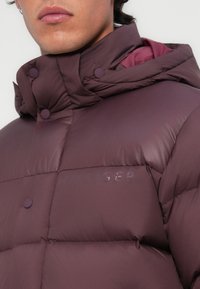 Marsala puffer jakna s visokim ovratnikom, s pritiskom dugmadi, kviltanom teksturom i utisnutim brendom na prsima. Obložena unutrašnjost.