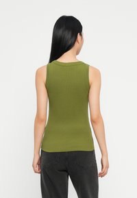 Vrouw met lang zwart haar, gekleed in een mouwloos olijfgroen topje en zwarte jeans, staande met haar rug naar de camera tegen een witte achtergrond.