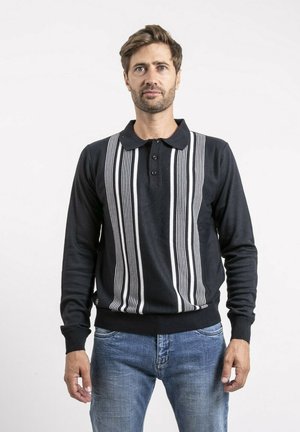 Homme aux cheveux châtain clair portant un pull polo noir à manches longues avec des rayures verticales blanches et un jean bleu, debout devant un fond blanc.