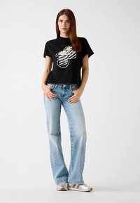 Black cropped t-shirt with graphic design, paired with light blue straight-leg jeans. Model wearing silver sneakers, hands on hips.