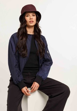 Giacca bomber navy con polsini a coste, top nero e pantaloni larghi neri. Il modello indossa un cappello a secchiello bordeaux. Texture liscia, design casual.