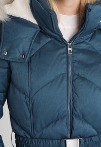 Polstret teal-jakke med høj krave, shearling-foring, lynlås lukning og elastisk talje. Har trykknapper og quiltet mønster.