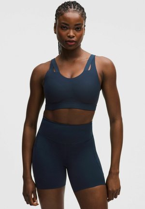 Athletic woman with braided hair wearing a navy sports bra and matching high-waisted workout shorts, standing against a plain background.