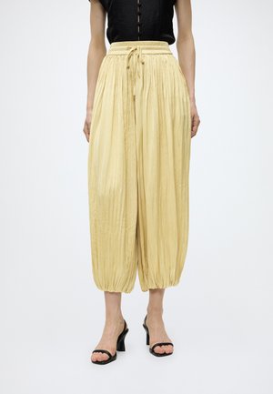 Femme portant un pantalon plissé ample jaune clair avec chevilles resserrées et taille à cordon, associé à des talons ouverts noirs et un haut noir.