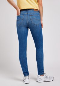 Personne portant un jean bleu ajusté et des baskets blanches, debout avec une main dans la poche arrière devant un fond uni.