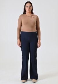 Beige langlopende top met een knoopdetail op de schouder, gecombineerd met marineblauwe op maat gemaakte broek. Model staat voor een neutrale achtergrond.