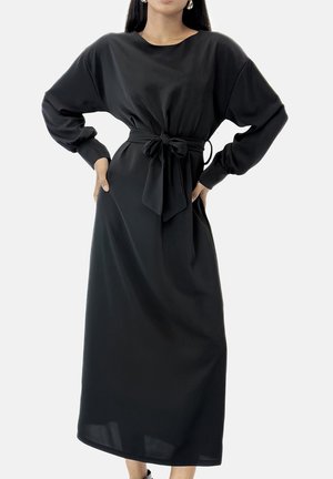 Femme portant une longue robe noire avec des manches ballon et un nœud noué à la taille, debout les mains sur les hanches.