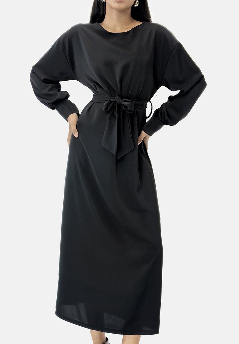 Femme portant une longue robe noire avec des manches ballon et un nœud noué à la taille, debout les mains sur les hanches.