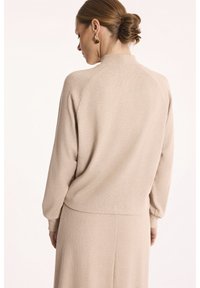 Beżowy sweter prążkowany z wysokim kołnierzem i długimi rękawami, z okrągłym dołem i luźnym krojem, noszony z pasującą spódnicą.